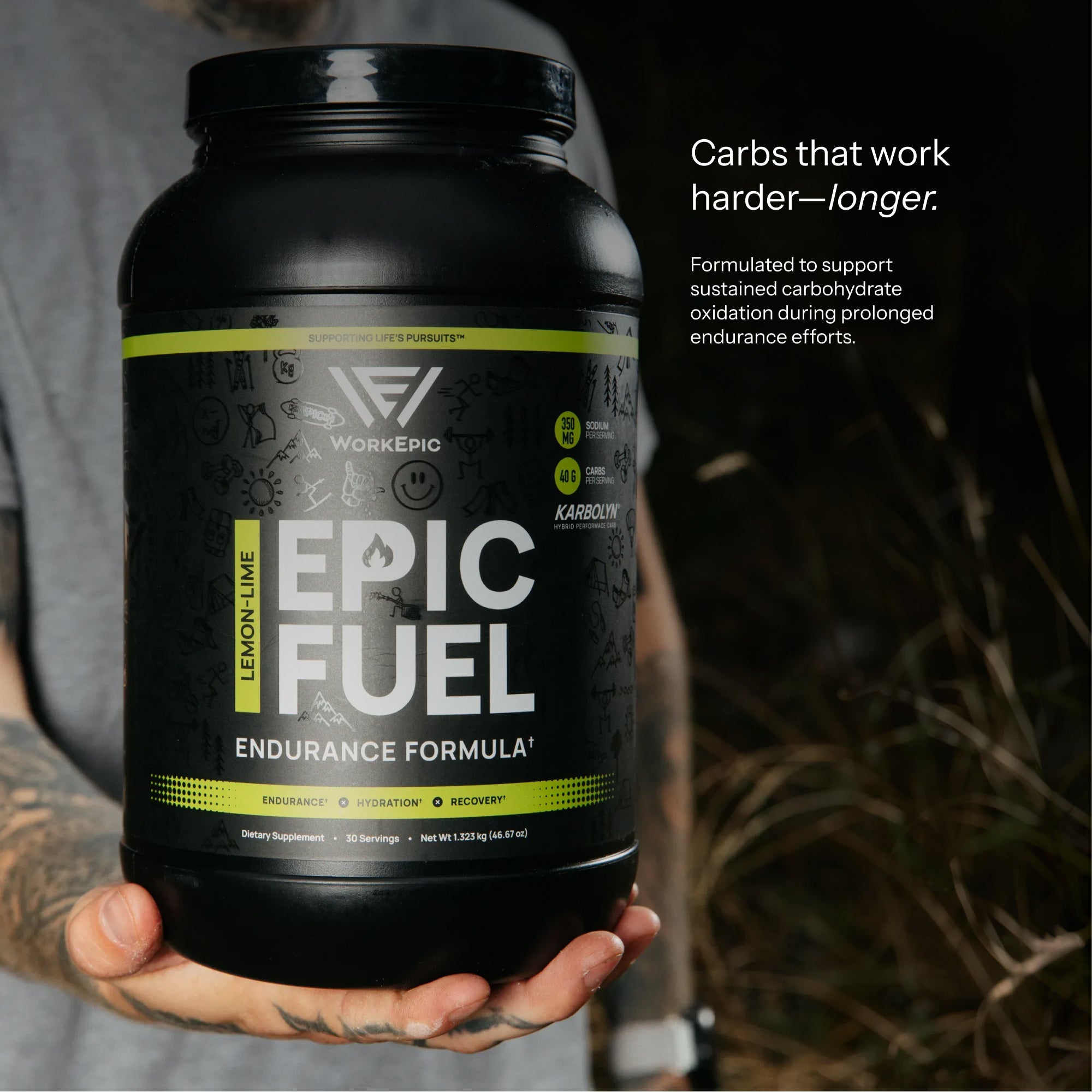 A person holds a large black tub of WorkEpic Epic Fuel Endurance Formula in Lemon Lime flavor. The background is a dark, blurred outdoor setting with natural light highlighting the product and the person's tattooed hand.