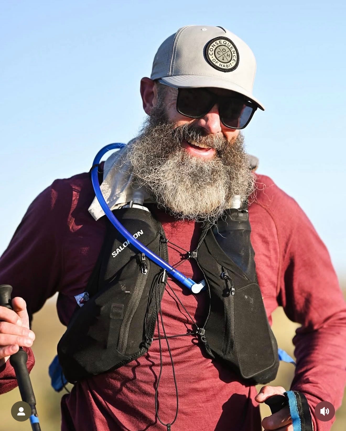 Man with a beard wearing sunglasses, a cap, and hydration gear outdoors.