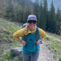 A woman hiking on a trail wearing a yellow and teal backpacking jacket, gray leggings, a blue and white patterned cap, and sunglasses, with a backpack on a grassy mountain path.