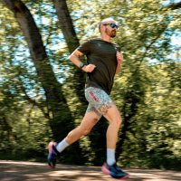 A man running outdoors on a wooded path, wearing a dark green t-shirt, patterned athletic shorts, white socks, and running shoes.