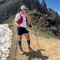 A male trail runner wearing a white t-shirt, black shorts, and running shoes is shown hiking with poles in a mountainous outdoor setting.
