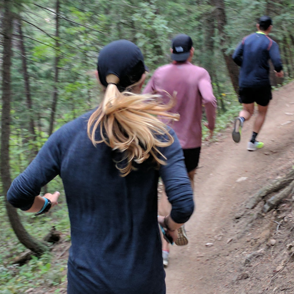 Three runners on a forest trail, with the foreground runner wearing a dark navy long-sleeve shirt and black cap seen from behind.
