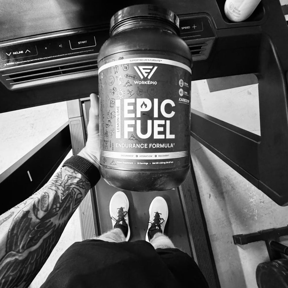 A person holding a large black tub of Workofo Epic Fuel Endurance Formula powder, standing on a treadmill in a gym setting. The photo is in black and white, showing the person's tattooed arm, shoes, and the treadmill in the background.