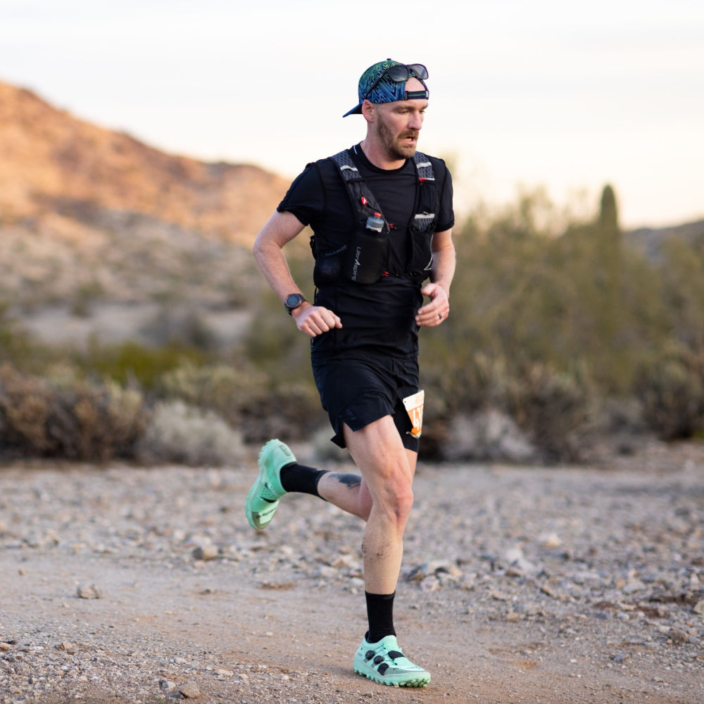 A male trail runner wearing a black t-shirt, black shorts, a black hydration pack, and a green cap is running on a rocky desert trail during golden hour.