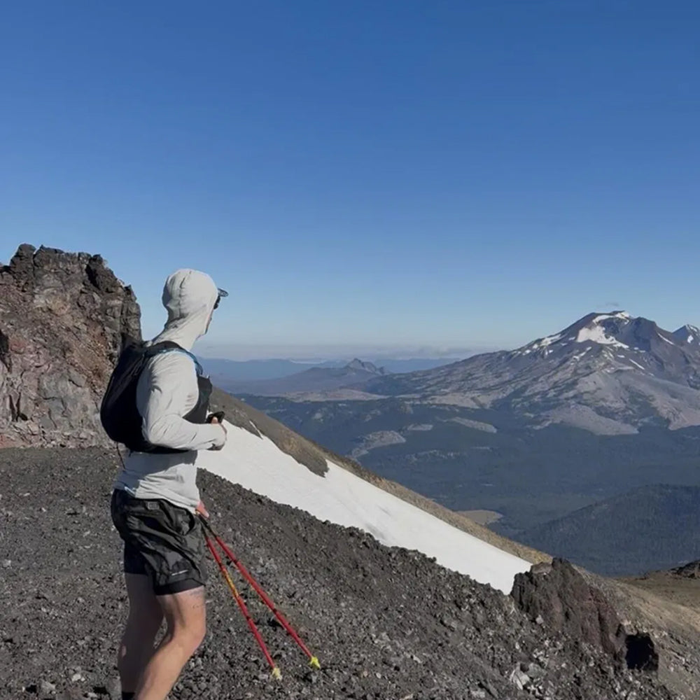 A hiker stands on a rocky mountain ridge wearing a light gray hooded long-sleeve top, black shorts, and a black backpack, holding red trekking poles and looking out over a snowy mountain landscape under a clear blue sky.